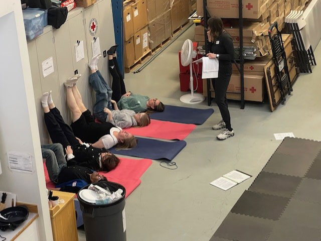Five people lie on mats with legs against a wall in a storage room, led by a person reading from notes.