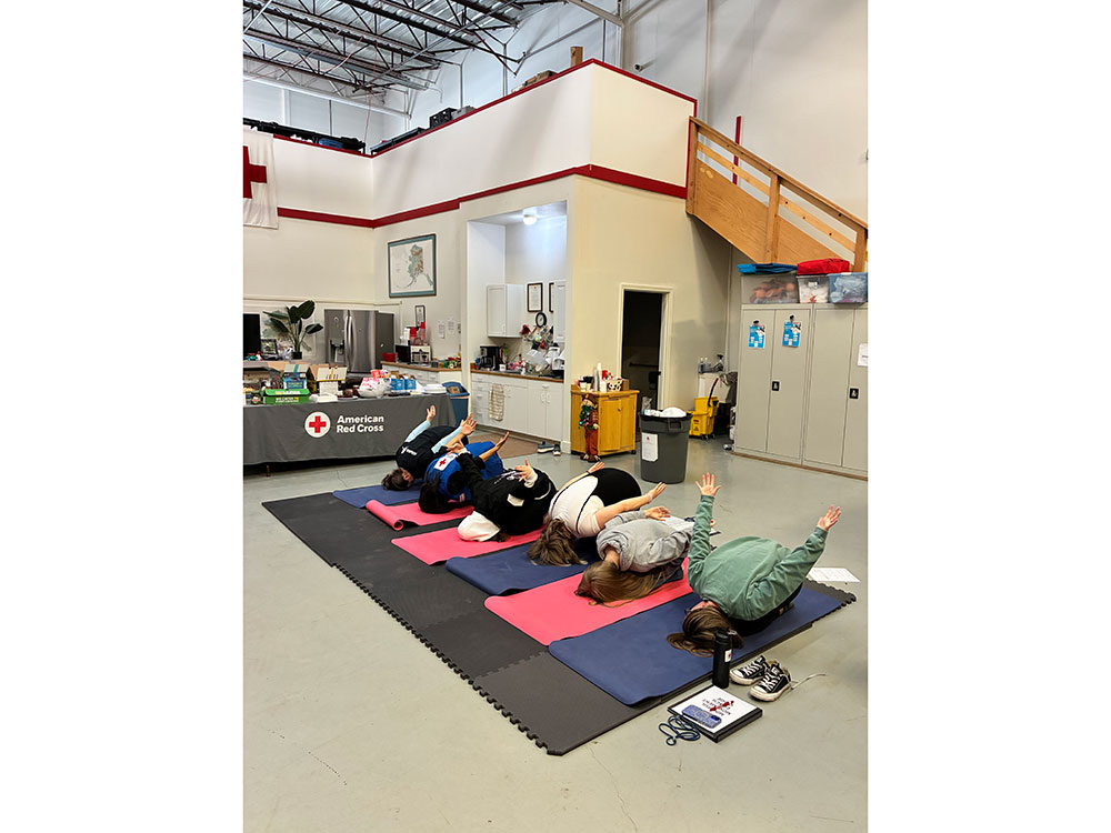 Five people are lying on colored mats in a yoga pose, extending arms upward. They're inside a room with American Red Cross signage visible.