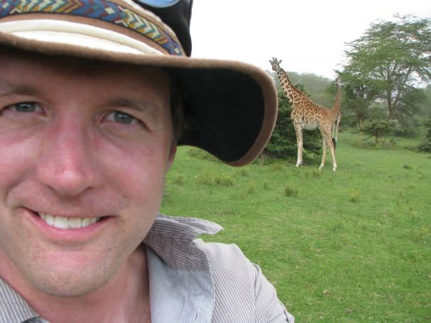 Bryce Sitter selfie while in Kenya with a two Giraffes in the background with one eating leaves from a tree.