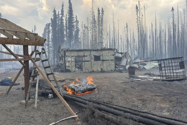 fire damage to a house with flames still burning and smoke filled air with tall trees in the background.