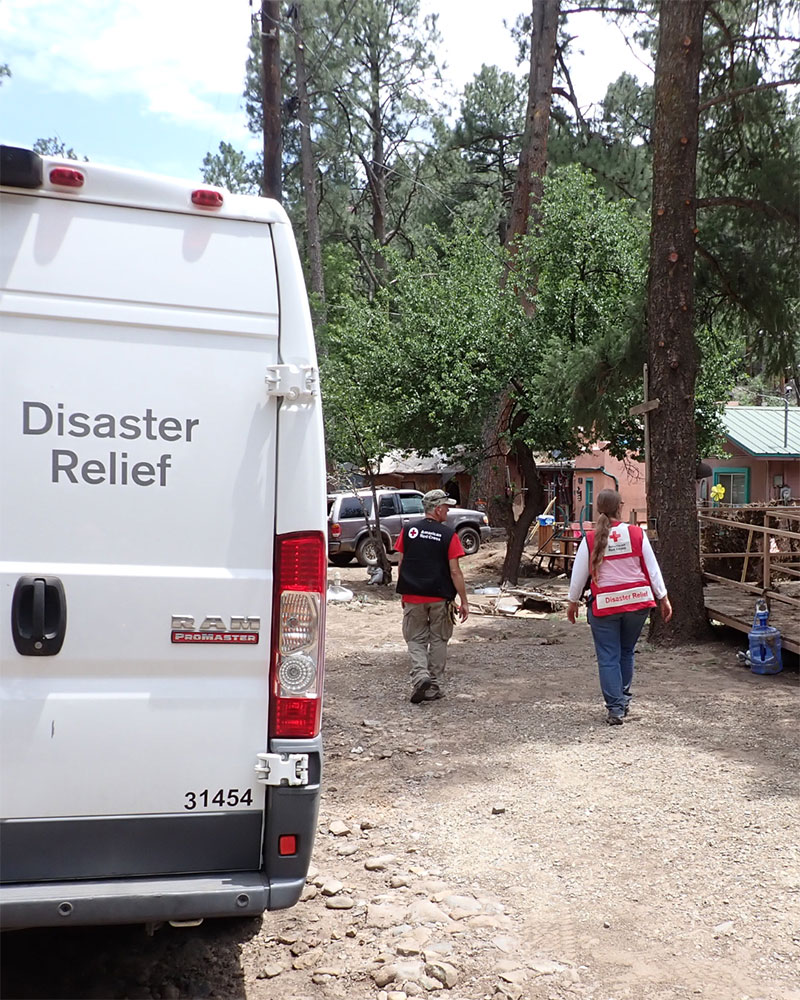 Red Cross volunteer Sofia Theard walking next to another volunteer towards houses that are surrounded by forest trees.