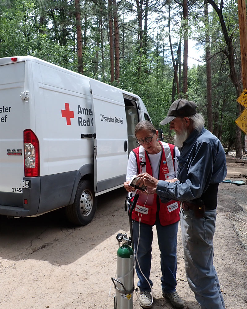 Red Cross volunteer Sofia Theard helping man with oxygen tank with red cross van and forest in the background.
