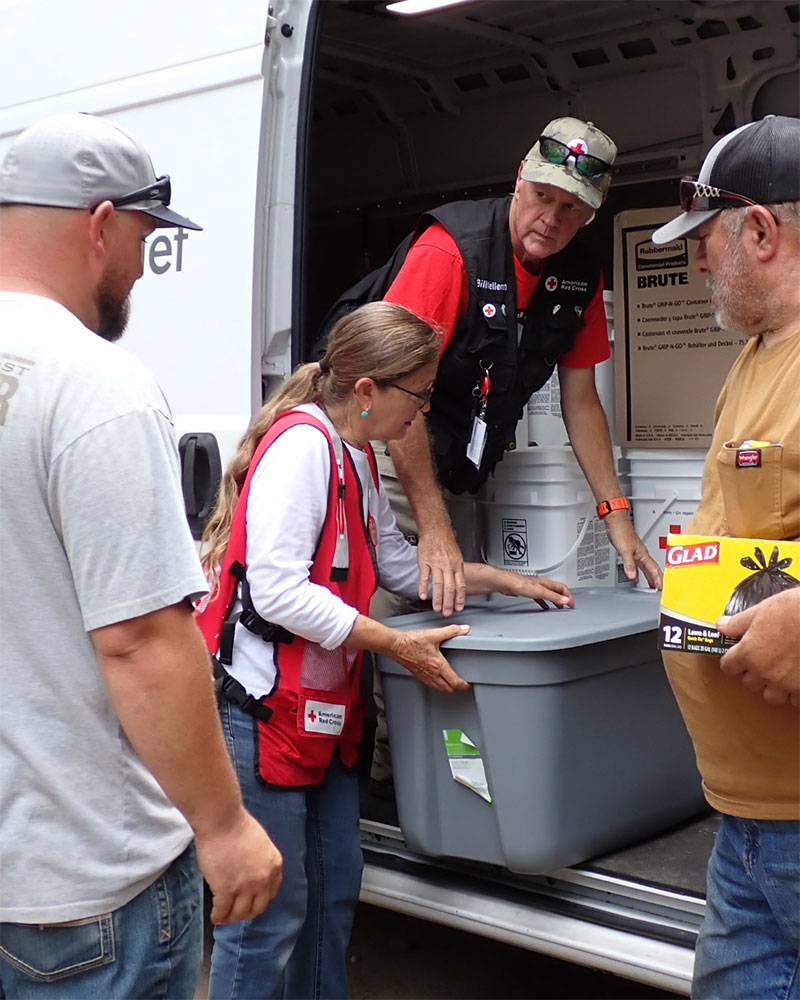 Red Cross volunteer Sofia Theard and another volunteer unloading plastic bins from a red cross van with two people standinig next to them.