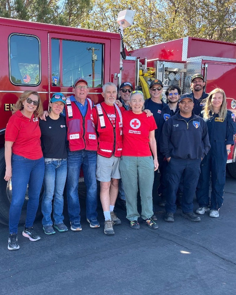 A group of smiling volunteers and firefighters stand in front of a red fire truck. They wear casual clothing and uniforms.