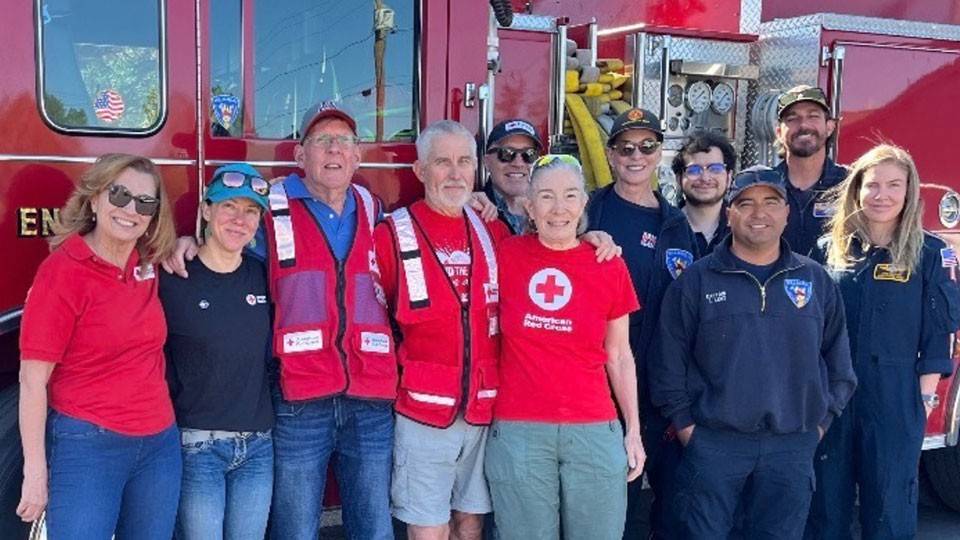 A group of smiling volunteers and firefighters stand in front of a red fire truck. They wear casual clothing and uniforms.