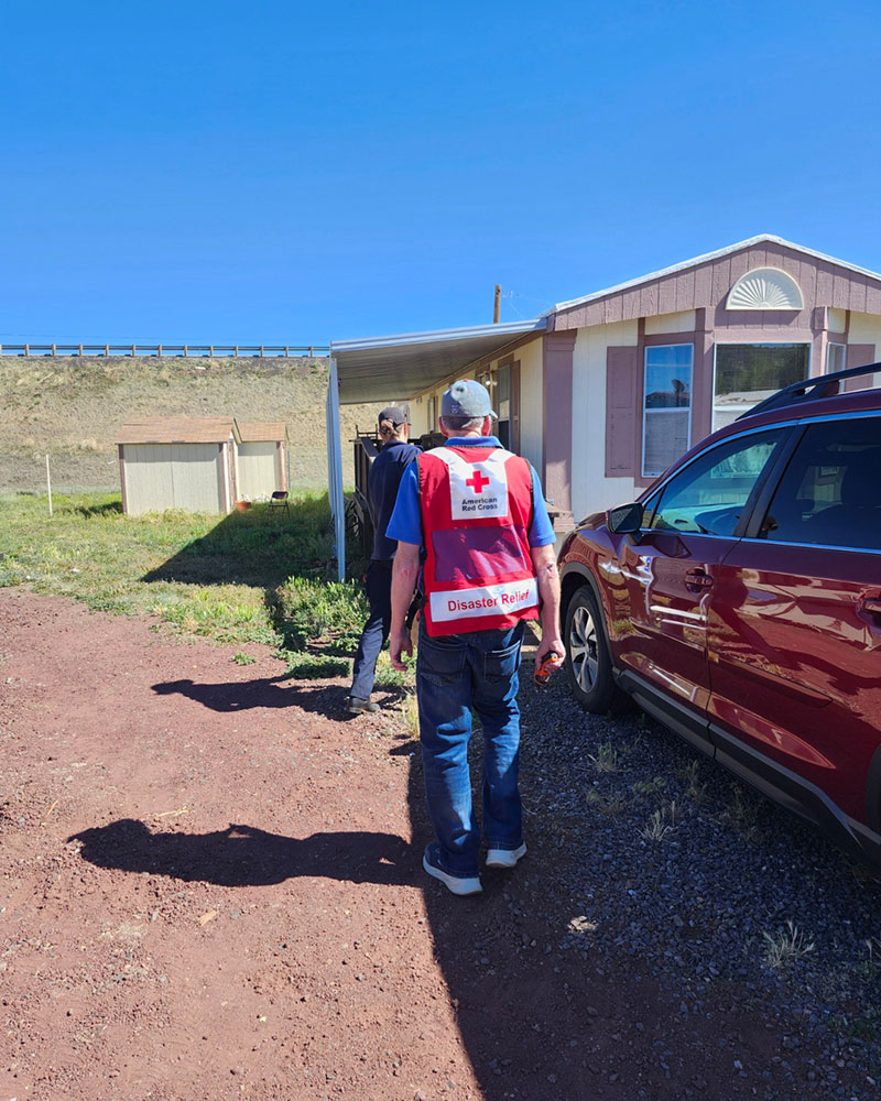A person in a Red Cross "Disaster Relief" vest walks towards a house with another individual, next to a red SUV under a clear blue sky.