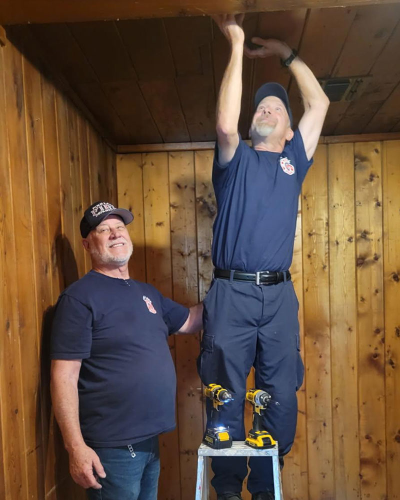 Two men in uniform are in a wooden paneled room. One stands on a ladder reaching up, while the other, smiling, supports the ladder with tools nearby.