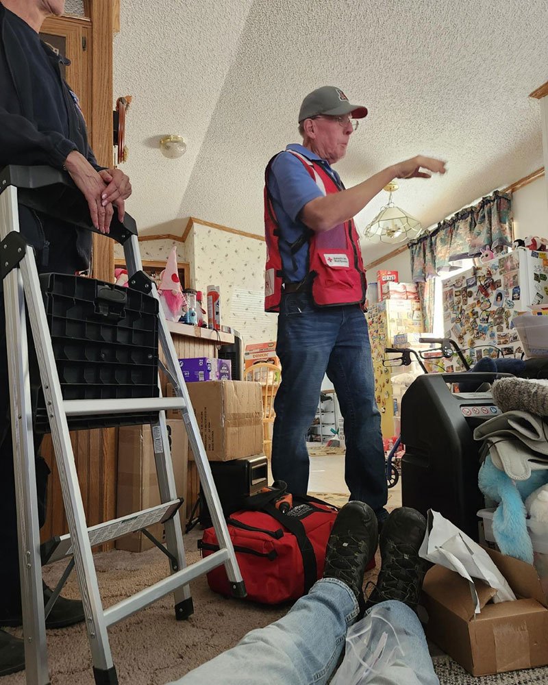 A man in a Red Cross vest speaks animatedly indoors, surrounded by cluttered boxes and a black cart. Another person stands near a folding ladder.