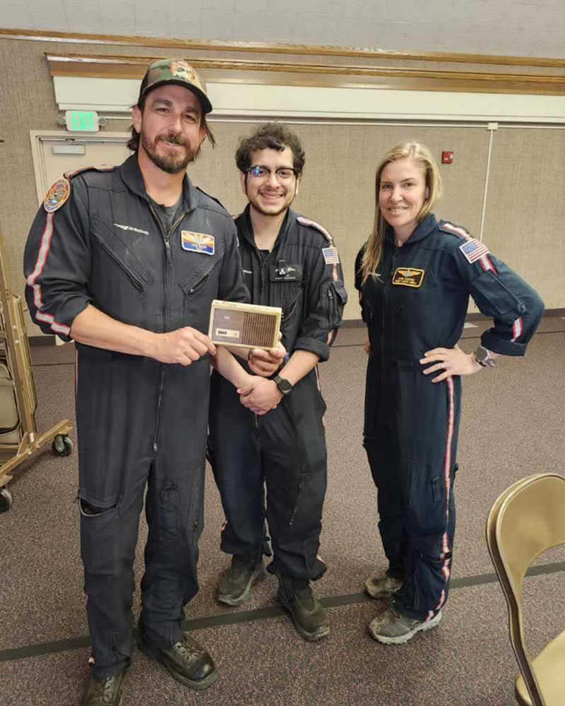 Three people in uniforms pose indoors; one holds a smoke alarm. They are smiling and stand in a relaxed, friendly manner.