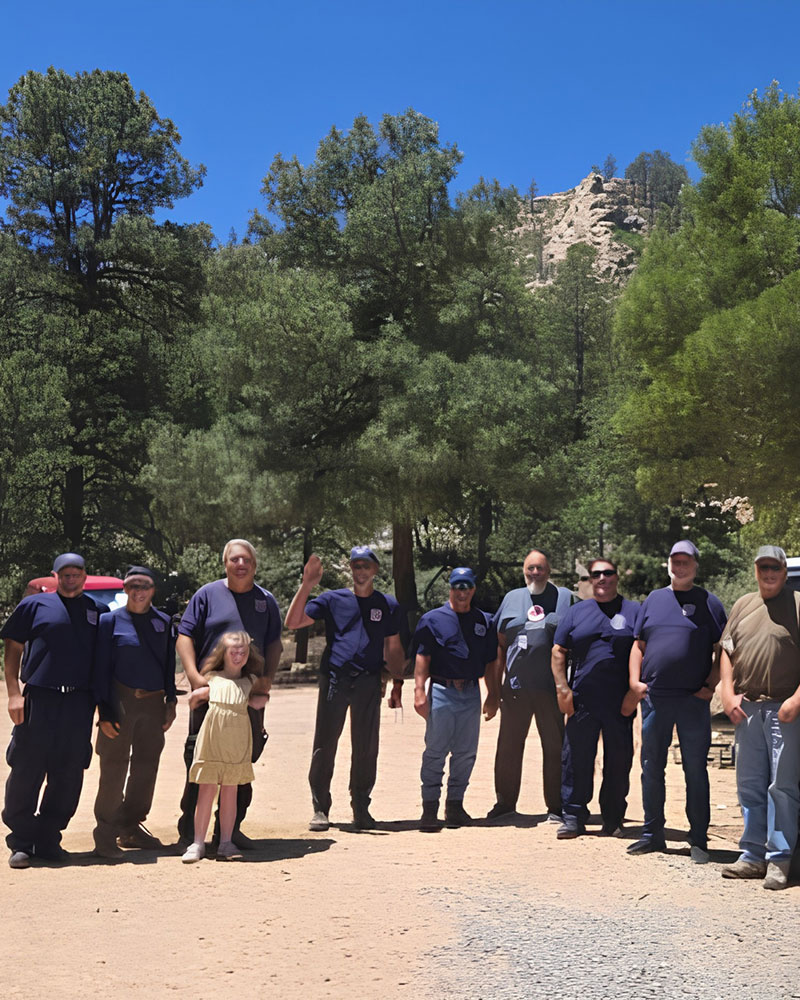 A group of people, mostly in matching blue shirts, stands outdoors on a sunny day. Tall pine trees and rocky hills form the scenic background.