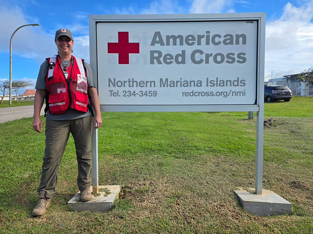 Red Cross volunteer Caroline Scudder standing in front of a an American Red Cross Northern Mariana Islands sign on the grass.