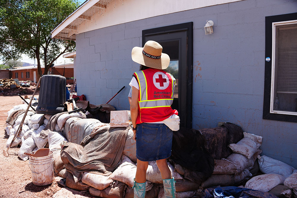 a Red Cross volunteer walking next to a house with sandbags stacked around.
