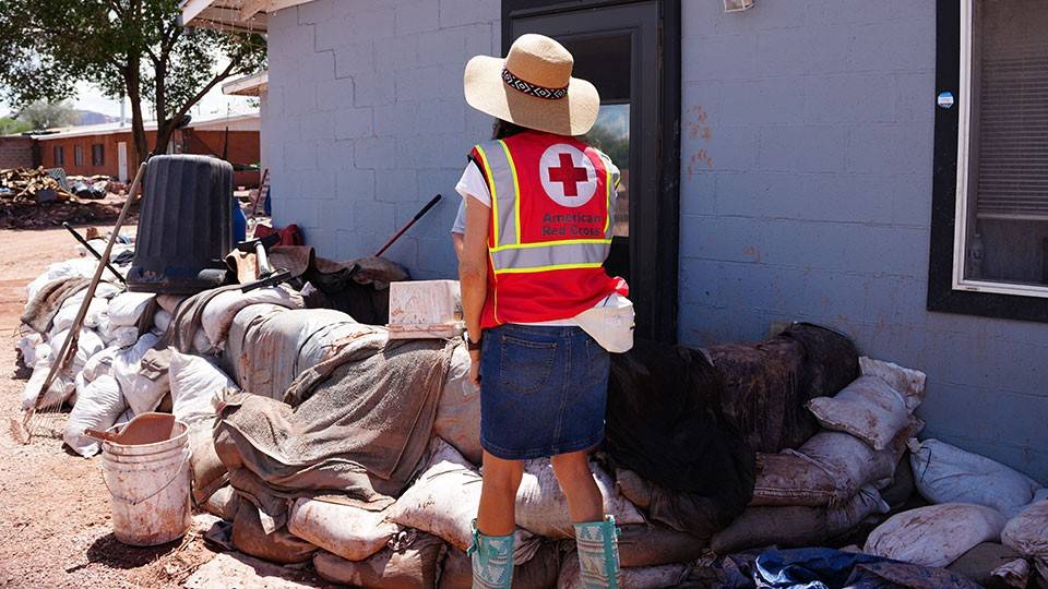a Red Cross volunteer walking next to a house with sandbags stacked around.