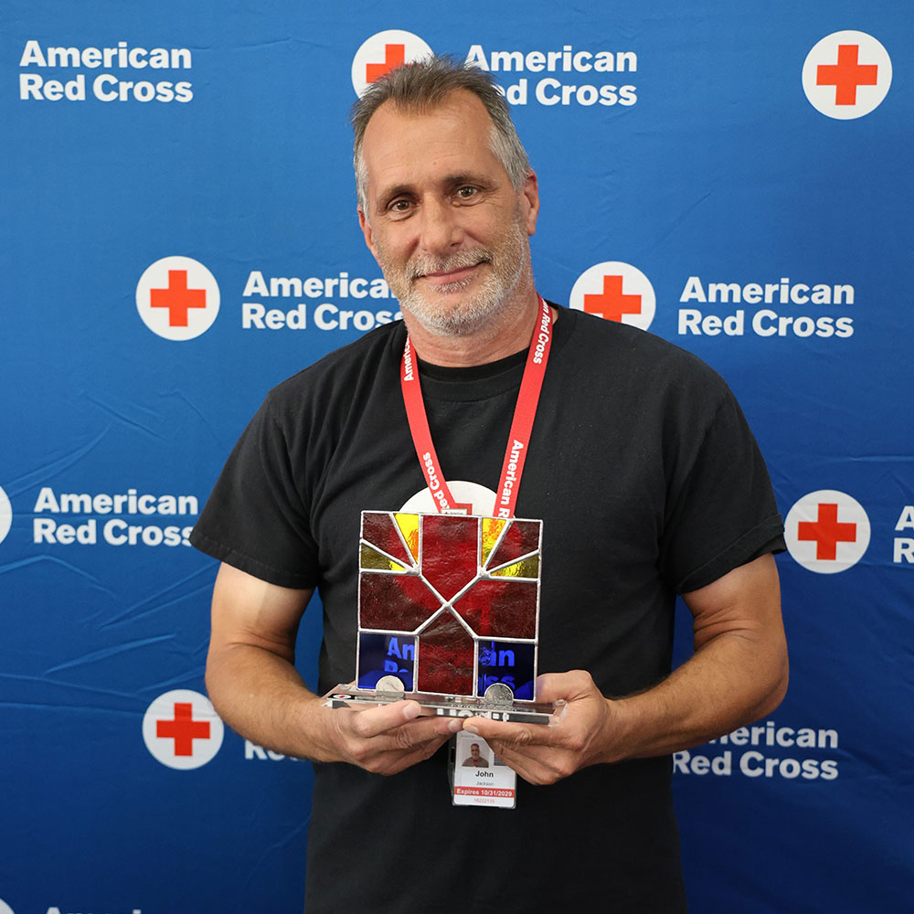 John Jackson holding a stained glass Red Cross award, smiling in front of a blue American Red Cross backdrop. He wears a lanyard and badge.