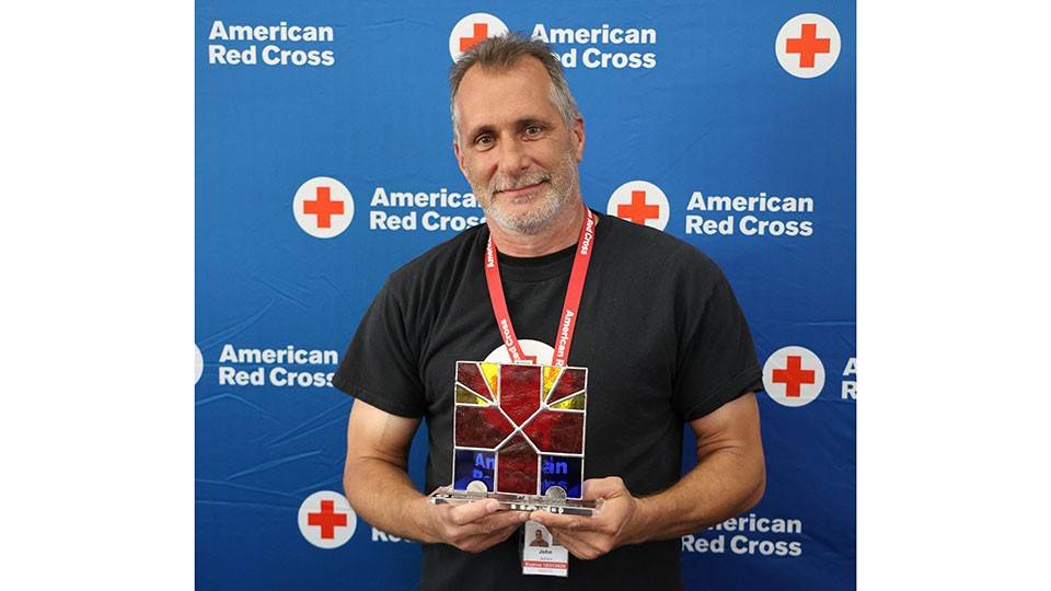 John Jackson holding a stained glass Red Cross award, smiling in front of a blue American Red Cross backdrop. He wears a lanyard and badge.