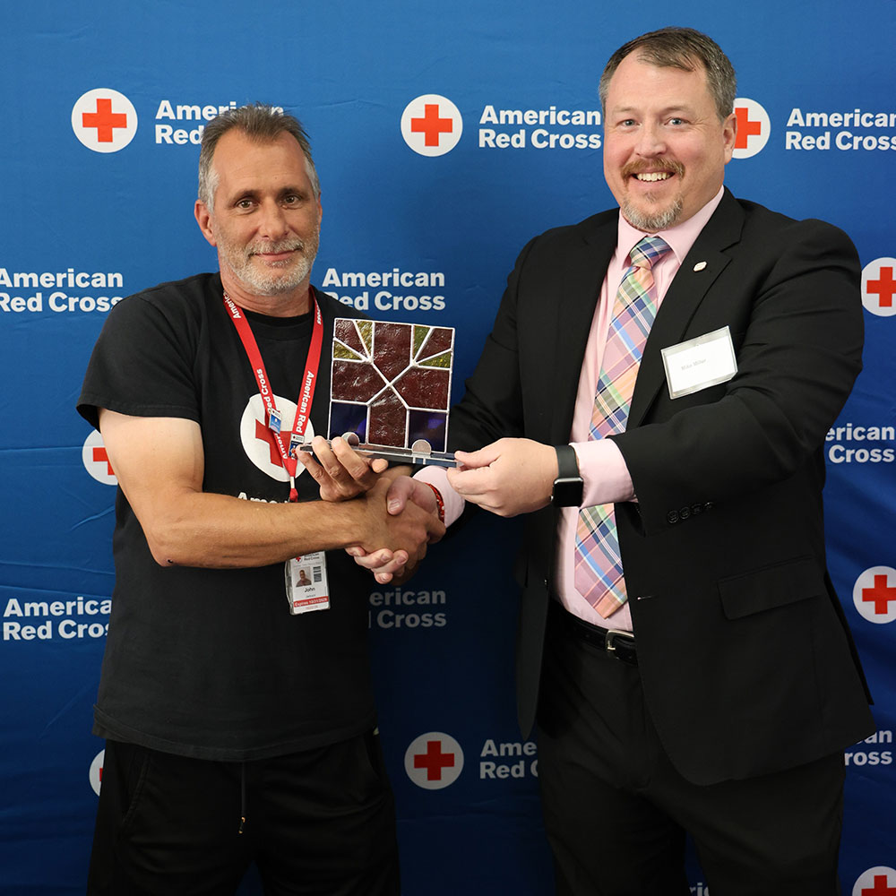 John Jackson and anoter man stand smiling, shaking hands while holding a stained glass Red Cross award. They are in front of an American Red Cross backdrop.