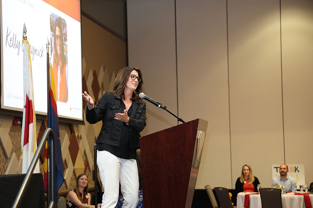 Kelly Waynick in a black jacket and white pants speaks at a podium with a microphone, smiling. A slideshow displays behind her. Audience members and flags are visible.
