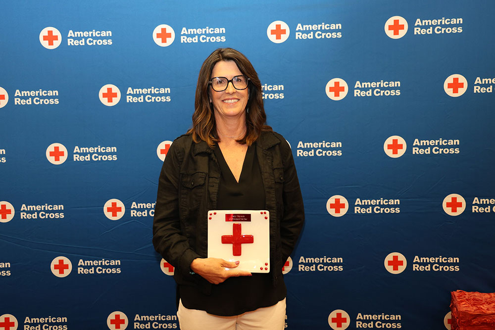 Kelly Waynick smiles while holding an award with a red cross on it. They stand in front of a blue American Red Cross backdrop.
