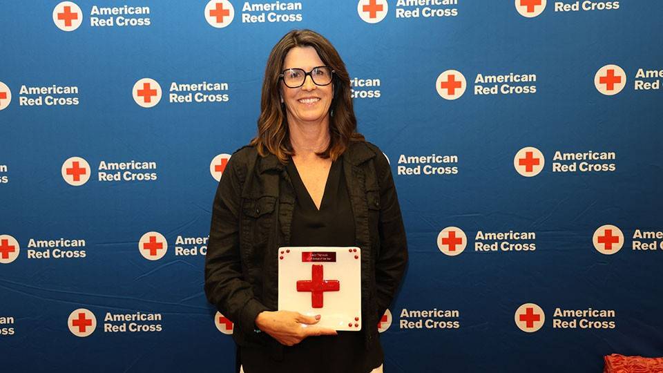 Kelly Waynick smiles while holding an award with a red cross on it. They stand in front of a blue American Red Cross backdrop.