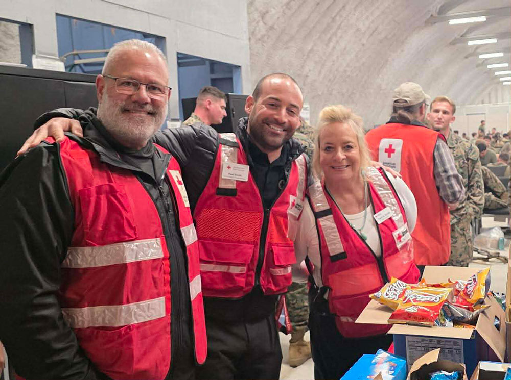 Tami and Paul Smith and Paul Smith Jr., wearing red cross volunteer safety vests, take a group photo while next to boxes with chips.