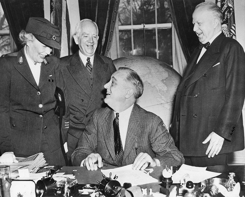 historical photograph from March 1943 showing President Franklin D. Roosevelt seated at his desk in the White House, smiling as he prepares to sign the Red Cross Month proclamation. Standing to his left is Mrs. Dwight F. Davis, in a Red Cross uniform and hat. Behind the President is Norman Davis, Chairman of the American Red Cross, and to the right stands an unidentified smiling man in a suit and bowtie. A large globe is visible in the background.