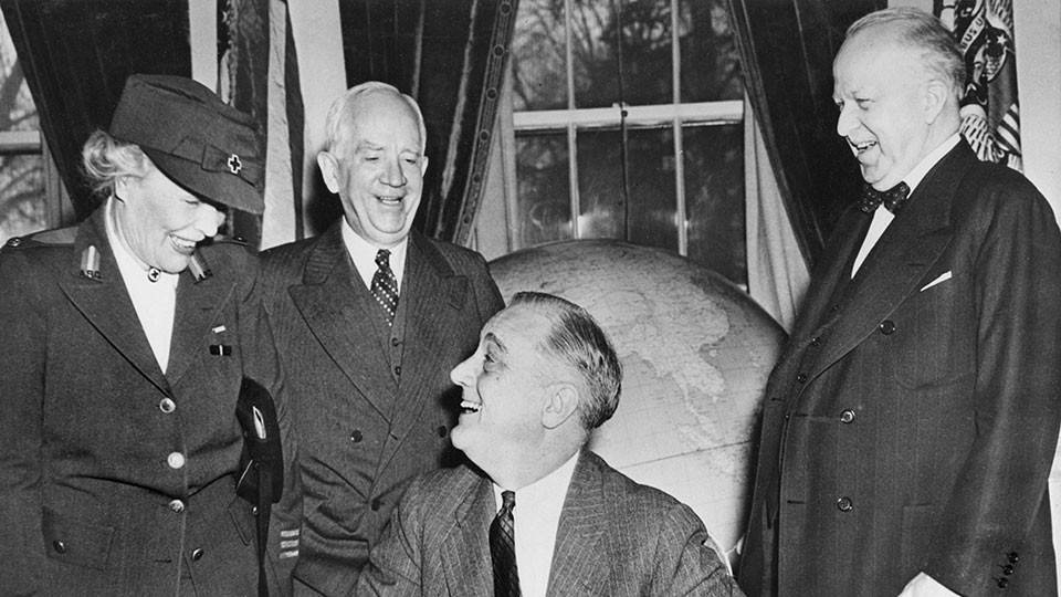 historical photograph from March 1943 showing President Franklin D. Roosevelt seated at his desk in the White House, smiling as he prepares to sign the Red Cross Month proclamation. Standing to his left is Mrs. Dwight F. Davis, in a Red Cross uniform and hat. Behind the President is Norman Davis, Chairman of the American Red Cross, and to the right stands an unidentified smiling man in a suit and bowtie. A large globe is visible in the background.
