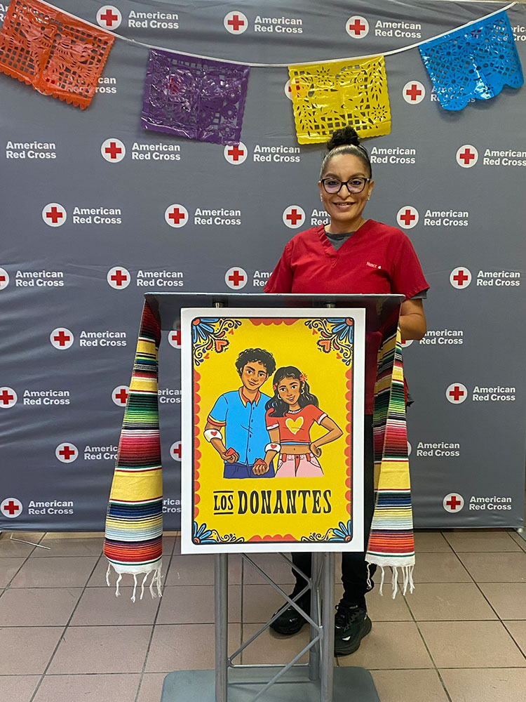 Reina Nunez in a Red Cross outfit standing behind the a podium with a Los Donantes poster.