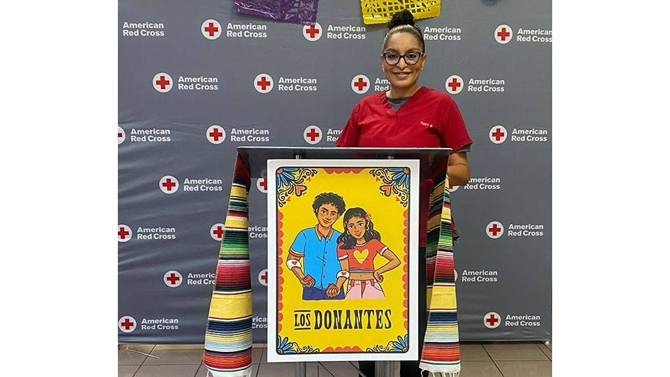 Reina Nunez in a Red Cross outfit standing behind the a podium with a Los Donantes poster.