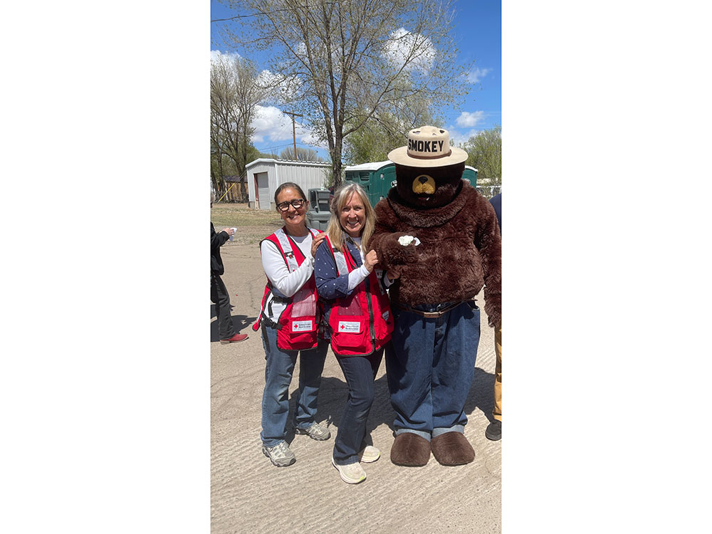 Two Red Cross volunteers pose beside Smokey the Bear mascot, smiling and enjoying a fun event together.