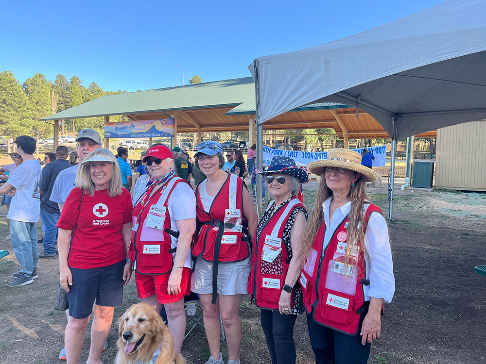 A group of red cross volunteers wearing red cross vests and hats stands beside a dog in a sunny outdoor setting.
