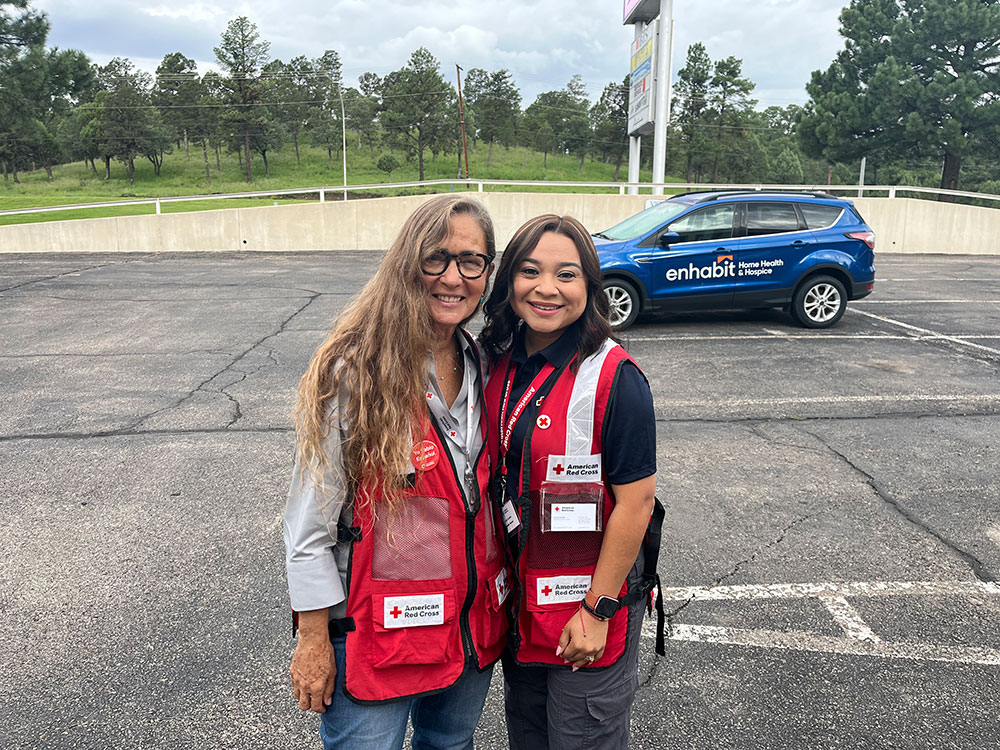 Two women in Red Cross vests smiling, standing in a parking lot with a blue "Enhabit Home Health & Hospice" car in the background.