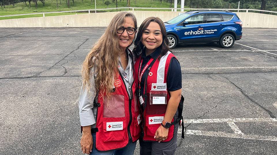 Two women in Red Cross vests smiling, standing in a parking lot with a blue "Enhabit Home Health & Hospice" car in the background.