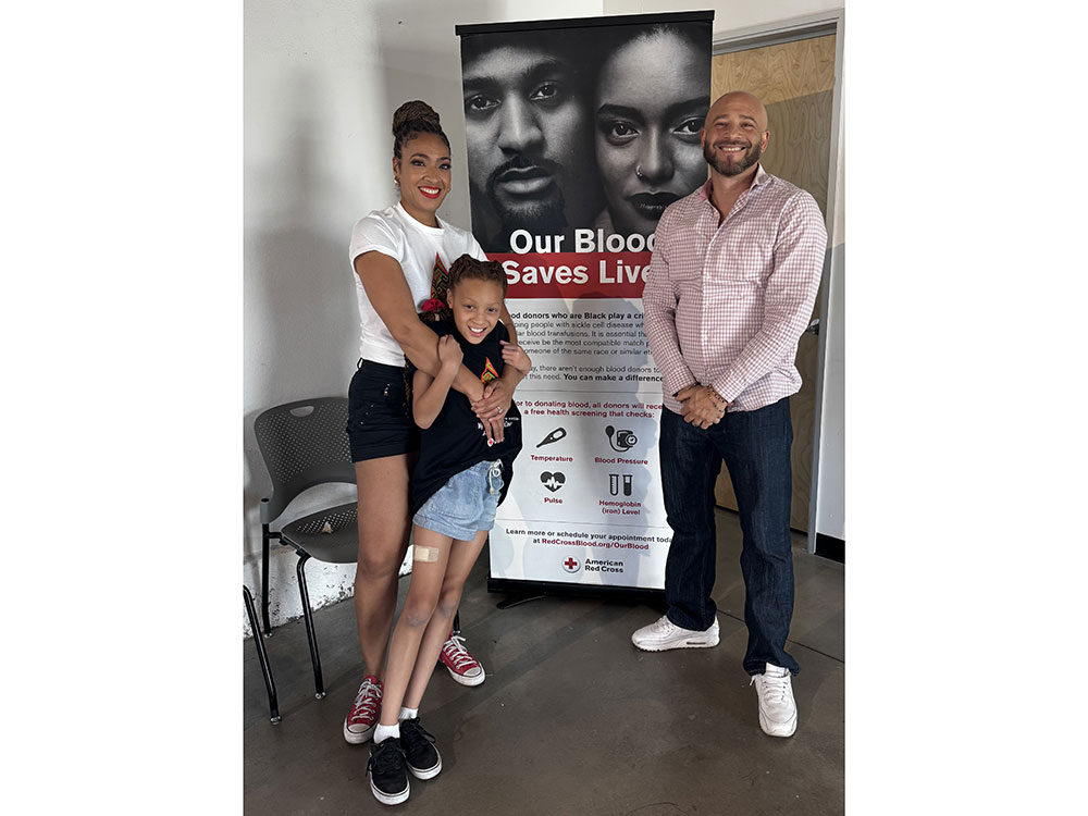 Dr. Aja Murphy, her daughter, and another person, stand smiling in front of a poster that reads "Our Blood Saves Lives."
