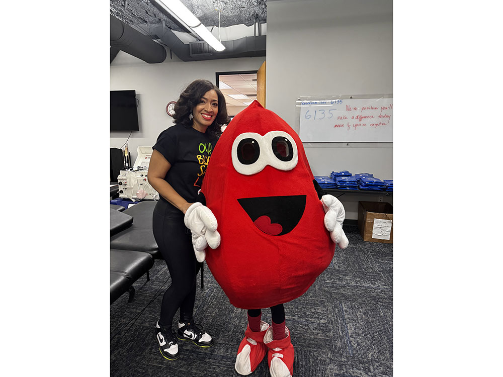Dr. Aja Murphy smiling in a black t-shirt stands beside a cheerful, red blood drop mascot.
