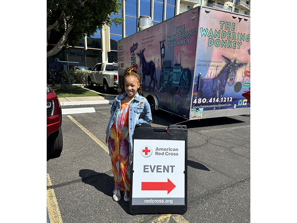 Dr. Aja Murphy's daughter stands next to an "American Red Cross Event" sign with a red arrow. Behind them is a colorful food truck labeled "The Wandering Donkey."