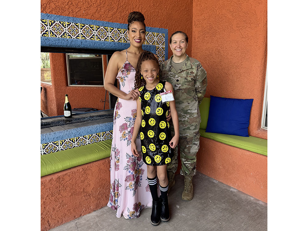 Dr. Aja Murphy in a floral dress, her daughter in a smiley face dress, and a person in military uniform stand smiling in front of a colorful tiled wall.