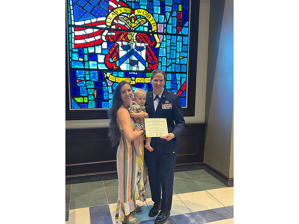 A person in a military uniform stands with a person holding a child, smiling and holding a certificate. A colorful stained glass window is behind them.