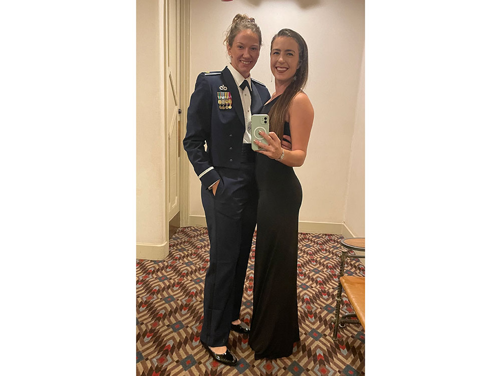 Two smiling individuals pose in a warmly lit room. One wears a military uniform adorned with medals, and the other is in a sleek black dress, conveying elegance.