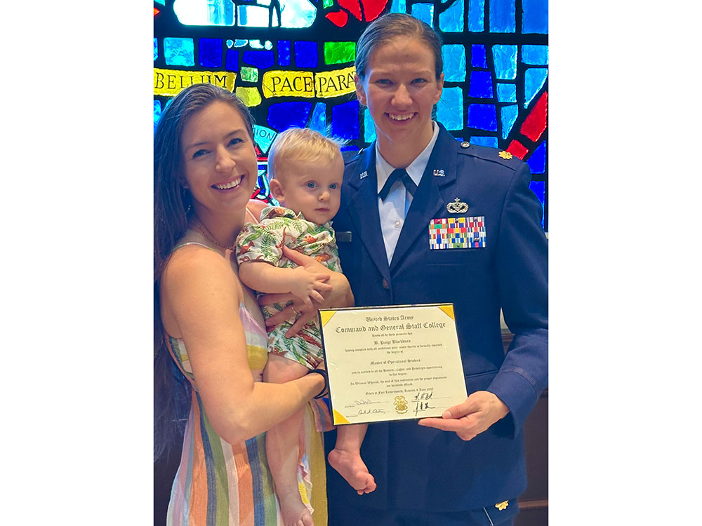 A person in a military uniform stands with a person holding a child, smiling and holding a certificate. A colorful stained glass window is behind them.