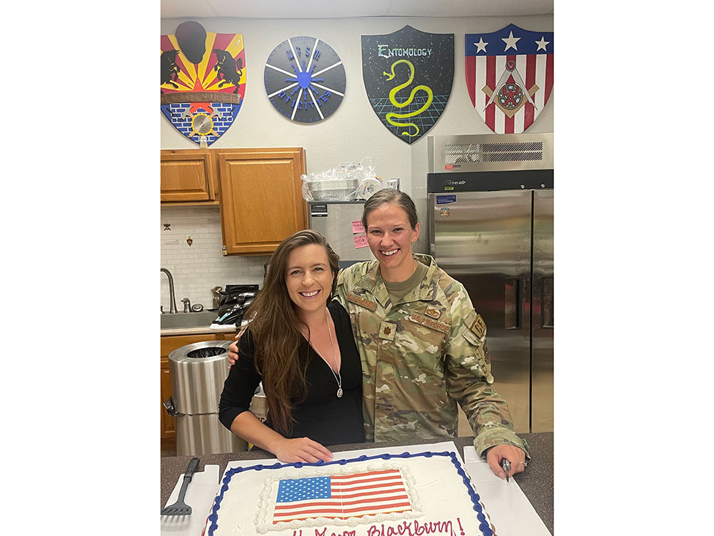 Two people smile in a kitchen, one in military uniform and the other in black. They stand by a cake with an American flag design.