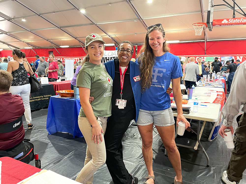 Three smiling people pose together indoors at a community event. They are surrounded by informational tables and attendees.
