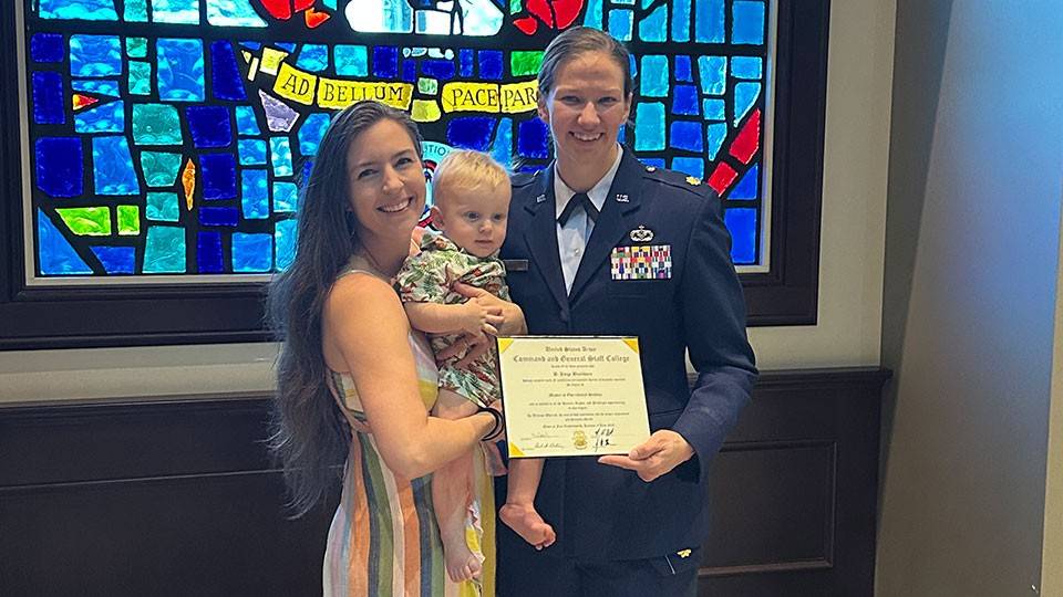 A person in a military uniform stands with a person holding a child, smiling and holding a certificate. A colorful stained glass window is behind them.