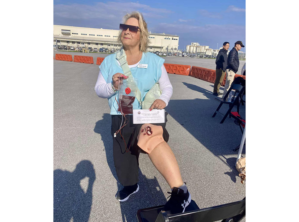 A woman with sunglasses holds a medical training device on her knee at an outdoor event. She's wearing a blue vest.