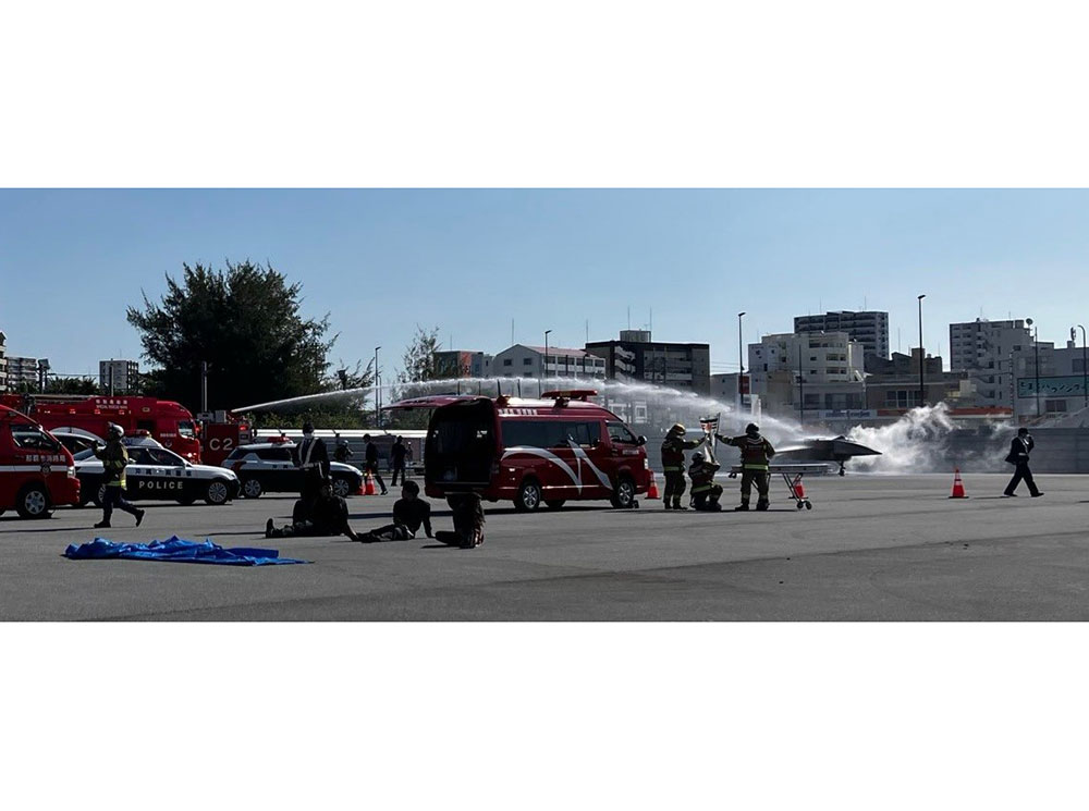 Emergency drill scene: firefighters spray water, red vehicles parked, people observing. Tall buildings in the background, clear sky overhead.