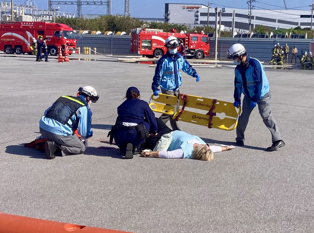 Rescue personnel in blue uniforms attend to a person lying on the ground during a drill. A stretcher is ready, fire trucks in the background.