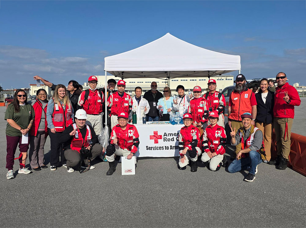 A group of smiling American Red Cross volunteers in red uniforms standing in front of a white tent on a clear day.