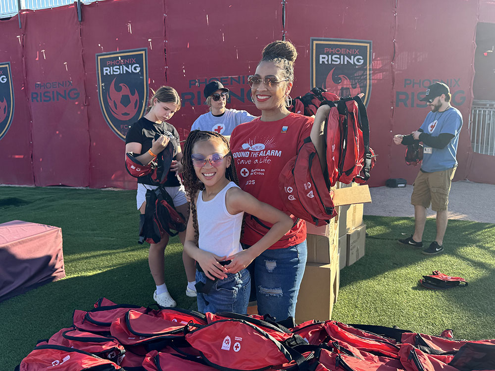 Dr. Aja Murphy and her daughter smile while holding Red Cross fanny packs at a community event. They are surrounded by volunteers and red banners in the background.