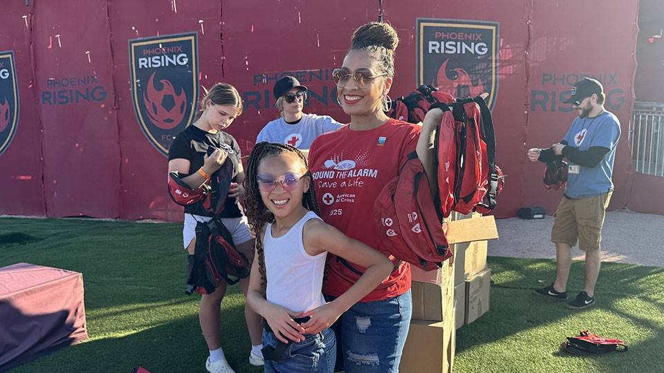 Dr. Aja Murphy and her daughter smile while holding Red Cross fanny packs at a community event. They are surrounded by volunteers and red banners in the background.