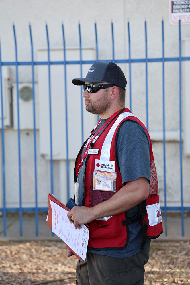 Scott Salzer outside wearing a hat, sunglasses, a Red Cross vest, and holding a clipboard.
