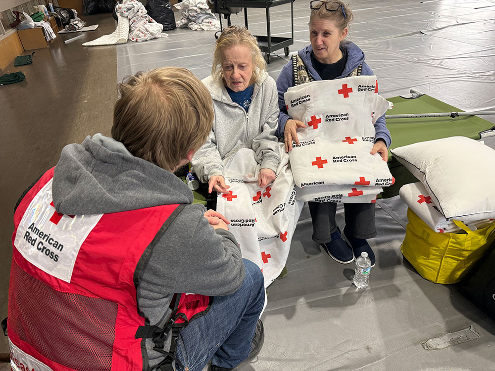 Beverly and Michele sitting on a cot while holding a Red Cross blanket, talk to a Red Cross volunteer at a shelter.